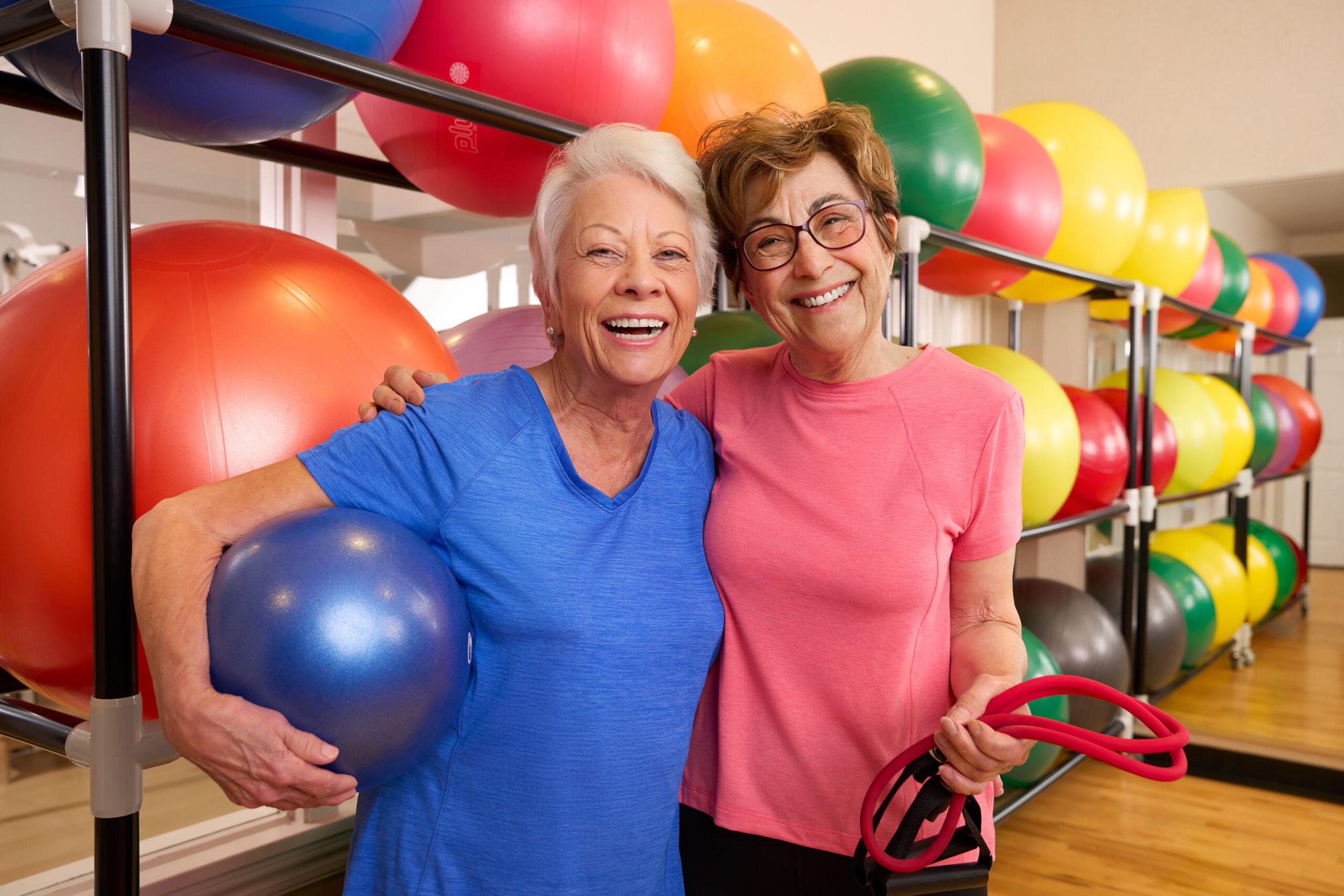 Two older women smile in front of a row of bright excercise balls.