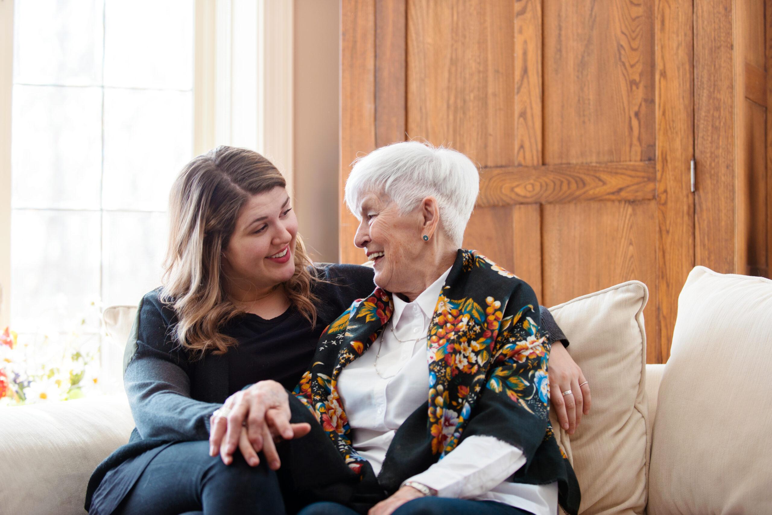 Adult daughter sits with her arm around her mother on the couch