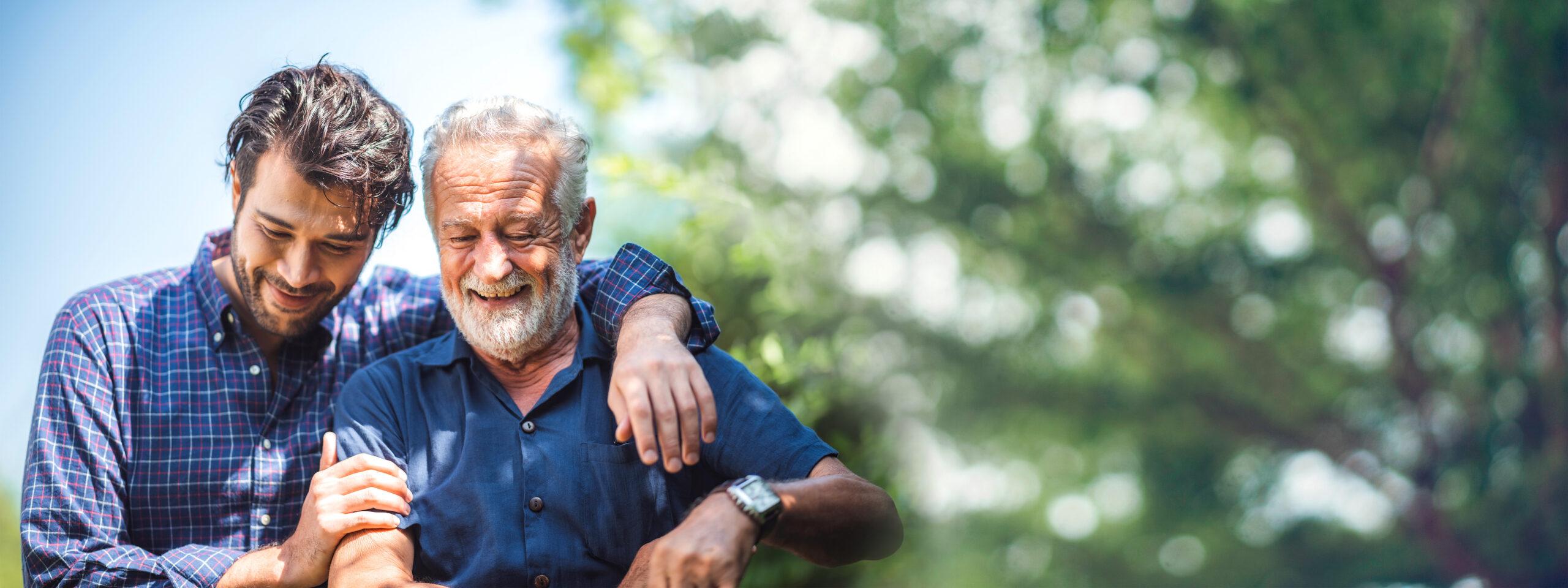 An adult son smiles with his arm around his father outdoors