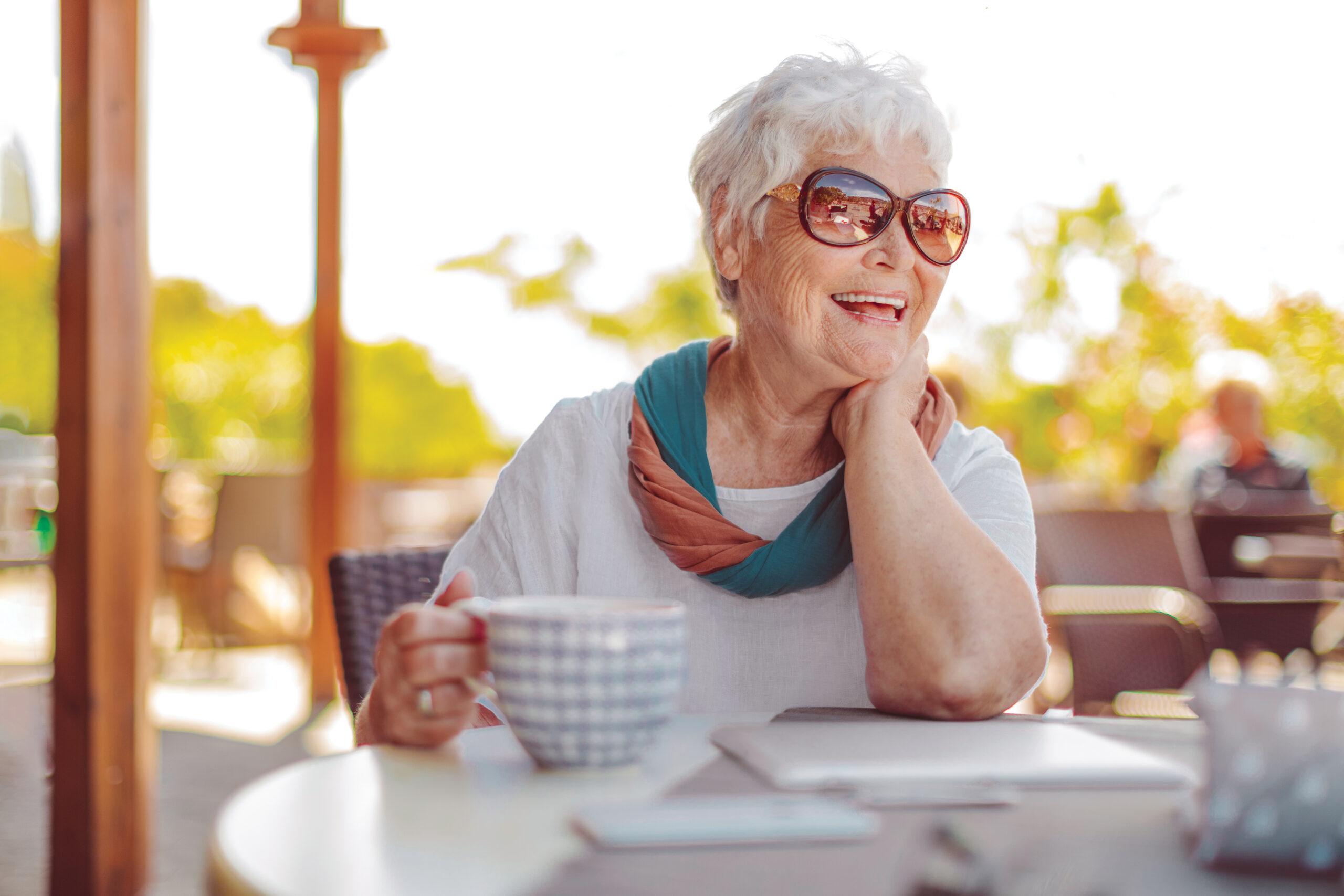 Older woman wearing sunglasses smiles and holds a hot drink