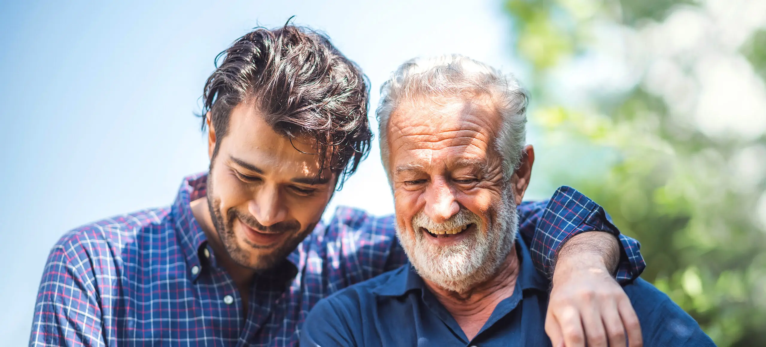 Adult son smiles with his arm around his father outdoors