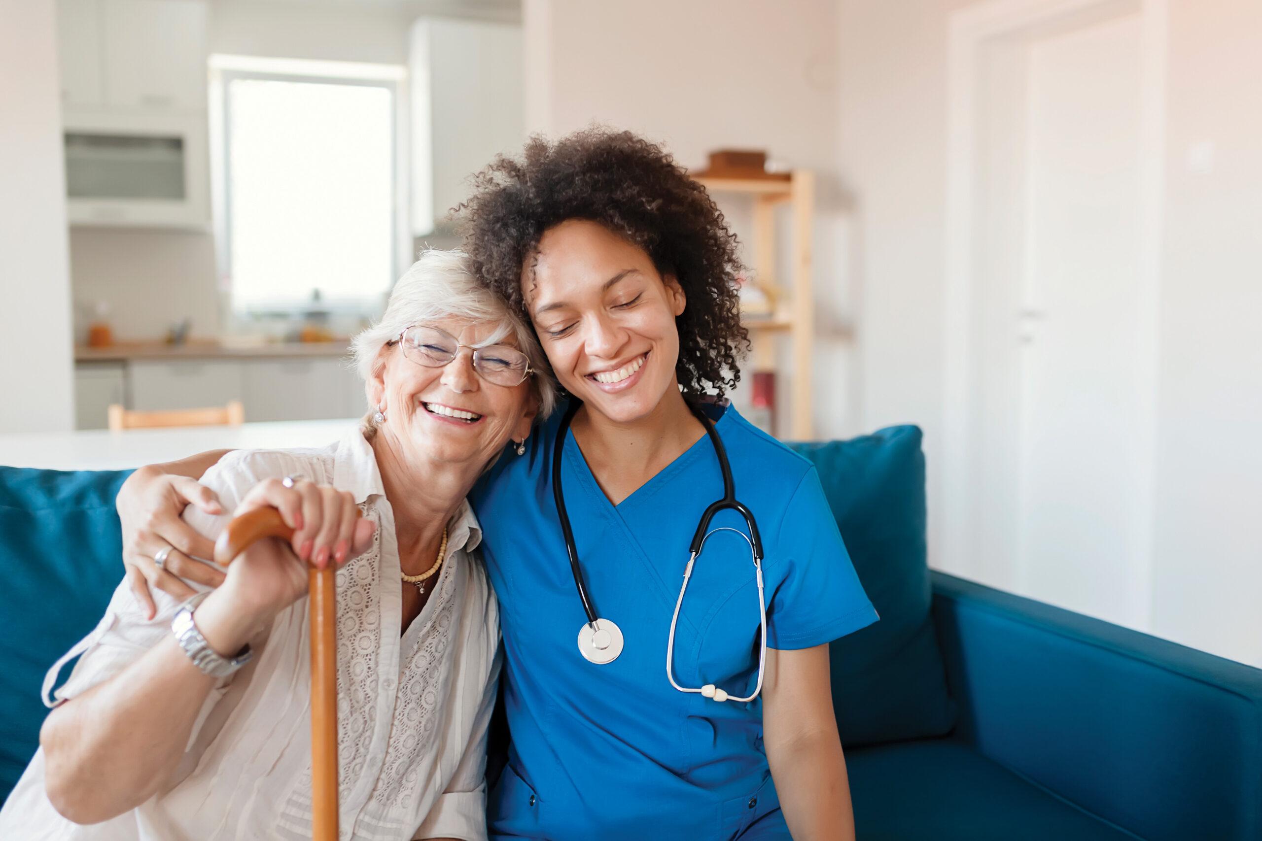 A caregiver sits with her arm wrapped around a smiling senior woman